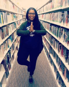 Amanda M. Leftwich is standing between two rows of bookshelves in a library. She is doing a yoga pose that inspires mindfulness.