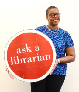 Aisha Conner-Gaten is standing wearing a blue shirt and against a white background. She is holding a round red sign that says the words Ask a Librarian, the text is white.