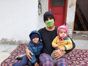 My husband (in a green face mask, black shirt and plaid pants) sitting in our small, cement courtyard holding out 11 month old on his knee and the 3 year old sits to the left smiling. Both are bundled for cold. My husband is Pathan and the children are both Pathan and white