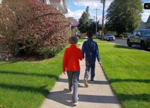 photo of 2 kids shown walking down a sunny street but from the back